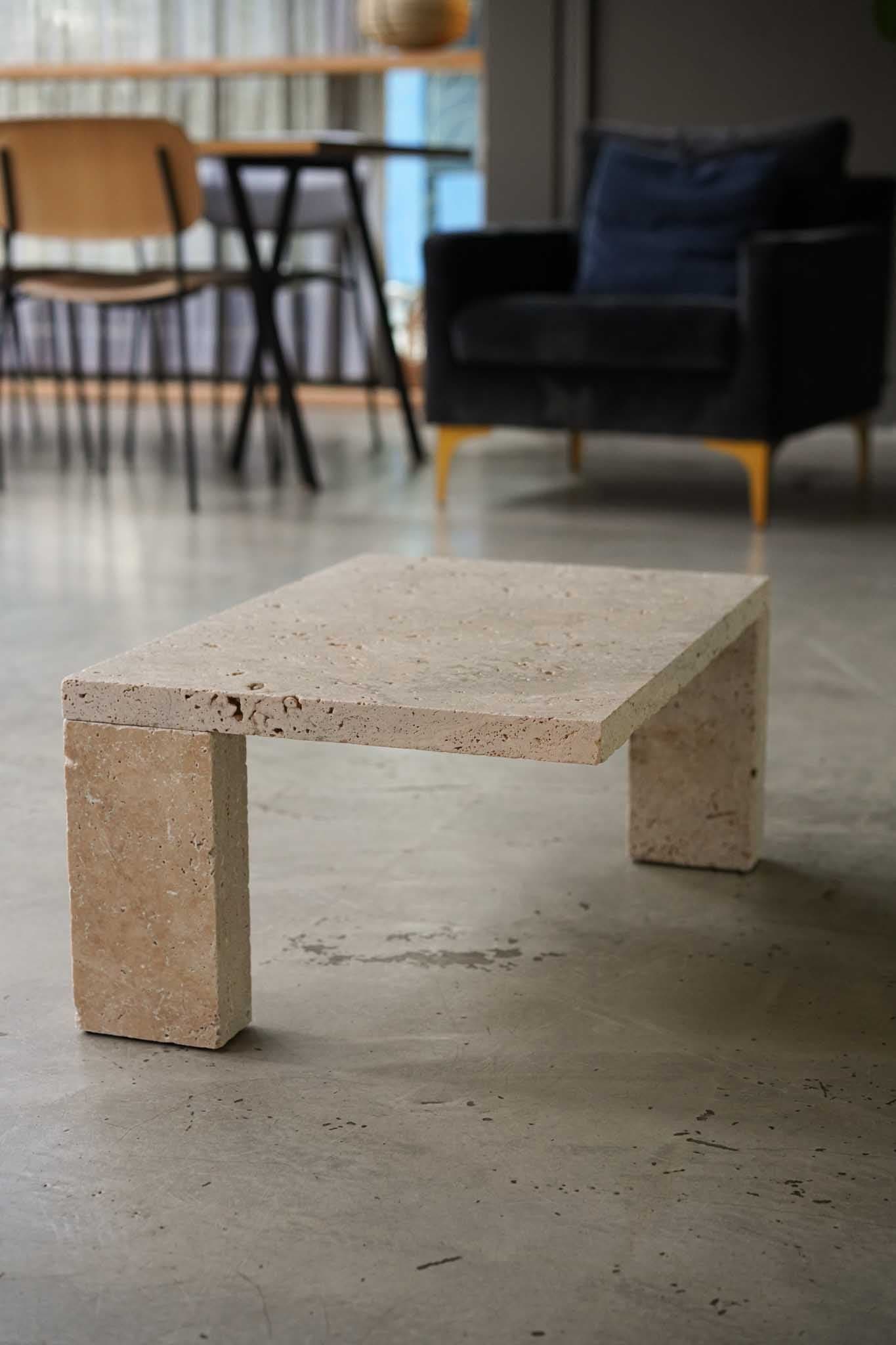 Sleek Base aus Travertin auf Betonboden, im Hintergrund ein dunkler Sessel